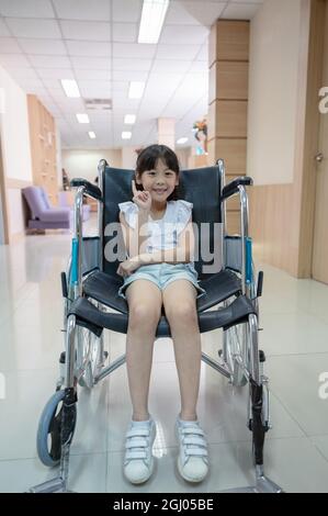 Fille asiatique mignonne souriante et levée de doigt en fauteuil roulant à l'hôpital Banque D'Images