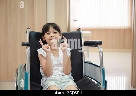 Fille asiatique mignonne souriante et levée de doigt en fauteuil roulant à l'hôpital Banque D'Images