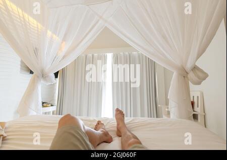 Homme étirant les jambes dans la moustiquaire sur le lit en vacances, lumière du soleil à travers le rideau blanc dans la chambre Banque D'Images