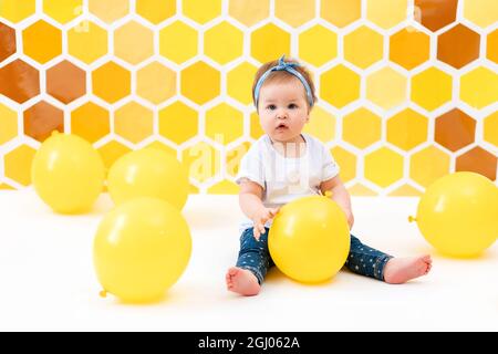 Le concept de la Journée mondiale de l'enfance. Une jeune fille est assise sur un sol blanc avec des ballons jaunes. À l'arrière-plan se trouvent des nids de miel jaunes. Banque D'Images