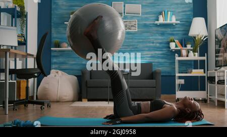 Femme noire à coupe près du corps tire-lait abdominal repose sur le sol avec un ballon suisse derrière les jambes pendant l'entraînement aérobie dans le salon. Adulte sportif portant des vêtements de sport bénéficiant d'un mode de vie sain Banque D'Images