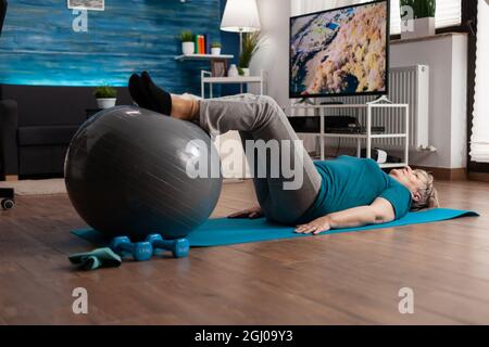 Femme âgée à la retraite pratiquant l'exercice des jambes en utilisant le ballon suisse assis sur le tapis de yoga dans le salon. Retraité retraité retraité dans le sport pratiquant abs cardio-travail à la santé musculaire du corps Banque D'Images