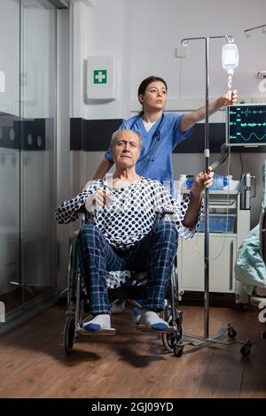 Médecin préparant un malade senior hospitalisé homme en fauteuil roulant, vérifiant iv goutte. Infirmière avec stéthoscope préparant le patient à l'examen. Oxymètre fixé au doigt de l'homme. Banque D'Images