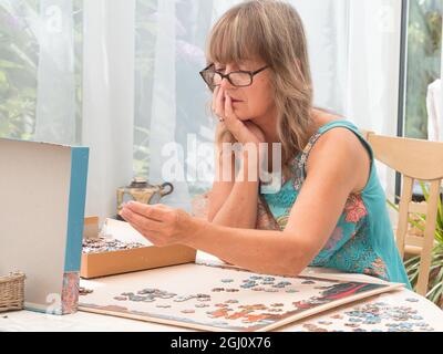 Une belle dame portant des verres se concentre dur avec le menton à la main regardant la boîte couvercle d'un puzzle de choisir la pièce à utiliser ensuite Banque D'Images