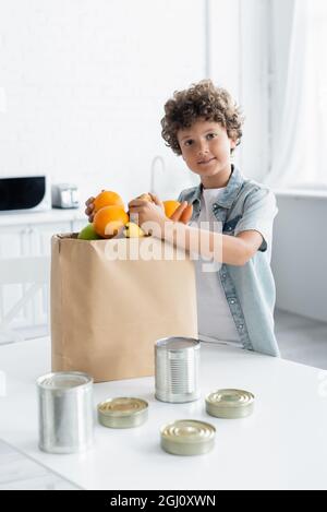 Garçon souriant tenant la nourriture près d'un sac en papier et de boîtes de conserve dans la cuisine Banque D'Images