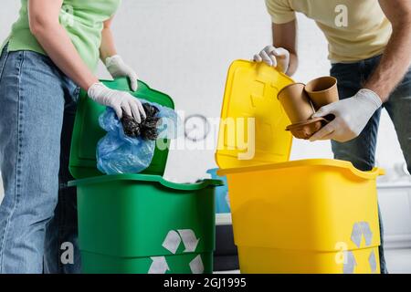 Vue rognée d'un couple en gants de latex triant les déchets dans les boîtes avec une affiche de recyclage à la maison Banque D'Images