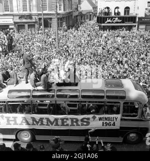 3 MAI 1964 Une partie des milliers d'Eastenders bordent la route à Stratford Broadway, dans l'est de Londres, comme l'équipe de football de West Ham United, vainqueurs de la finale de la coupe FA d'hier à Wembley, a défilé dans les rues de West et East Ham avec le trophée convoité. Bobby Moore, capitaine du West Ham, est fier de vous présenter le trophée. Derrière lui se trouvent Ken Brown (à droite) et Johnny Byrne (troisième de droite). Tant de gens étaient bondés dans l'hôtel de ville de Stratford que le maire a dû appeler la réception officielle de l'équipe. La police craignait que si l'équipe quittait une fois son entraîneur, elle ne puisse pas le faire Banque D'Images