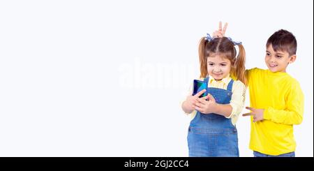 Le garçon souriant a mis deux doigts dans la forme de cornes sur la tête de la sœur et elle tient le téléphone, faisant selfie. Banque D'Images