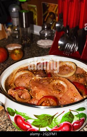 Mariné dans la sauce soja et la moutarde avec des oignons et des tomates coupés en rondelles, des tranches de saindoux de porc crues dans une grande casserole blanche sont sur la table dans la cuisine. À l'arrière-plan, tomates jaunes et rouges entières, sel, poivre et ustensiles de cuisine. Photo de haute qualité Banque D'Images