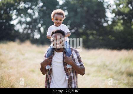 Beau homme africain en chemise à carreaux debout sur le terrain d'été et tenant son petit fils sur les épaules. Mignon garçon et son père souriant et regardant l'appareil photo à l'extérieur. Banque D'Images