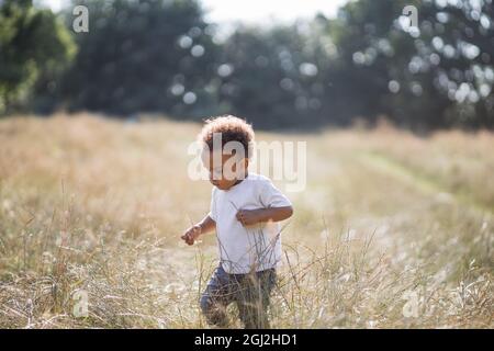 Petit garçon afro-américain jouant et courant sur le terrain parmi les herbes hautes. Modes de vie d'été et concept d'enfance insouciant. Banque D'Images