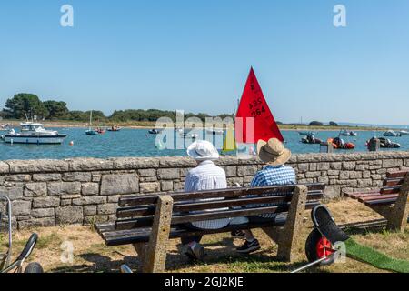 Couple senior assis sur un banc d'observation des bateaux et des dinghies naviguant à Keyhaven, Hampshire, Angleterre, Royaume-Uni Banque D'Images