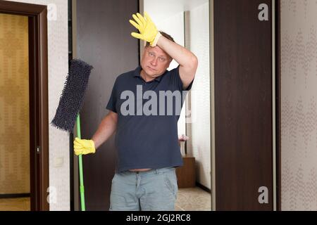 Un homme en gants de caoutchouc jaune avec une vadrouille dans les mains frotte son front de la fatigue après le nettoyage de l'appartement. Banque D'Images