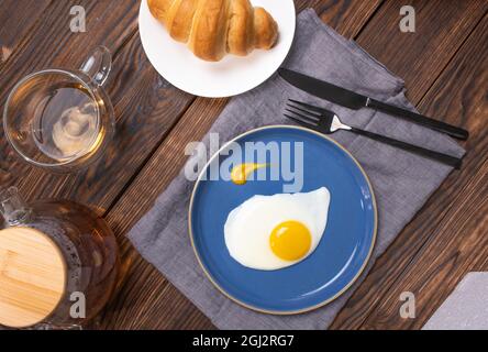 Œufs frits dans une assiette bleue sur une table en bois, petit déjeuner avec croissant et thé fraîchement préparé. Vue de dessus. Banque D'Images