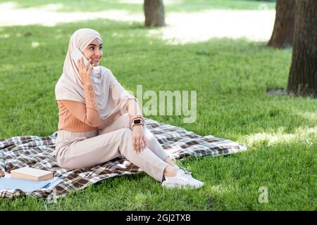 Jeune femme souriante du Moyen-Orient dans le hijab appelant par téléphone, détendez-vous dans le parc et profiter de la journée ensoleillée à l'extérieur. Jolie dame prend une pause de travail, communic Banque D'Images