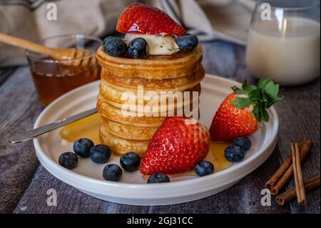 Vue rapprochée de la pile de crêpes avec bâton de miel, fraise, baie bleue et crème fouettée sur l'assiette, délicieux dessert avec lait pour le petit déjeuner. Banque D'Images