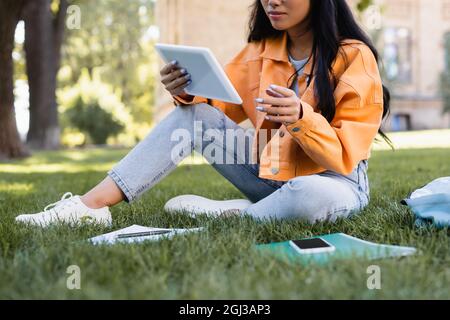 vue rognée de l'élève dans une veste orange à l'aide d'une tablette numérique sur l'herbe dans le parc Banque D'Images