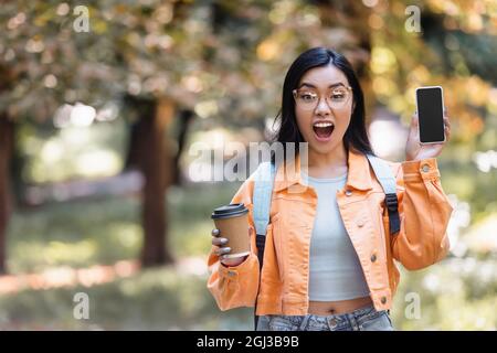 élève asiatique étonné avec café à emporter montrant téléphone mobile avec écran vide dans le parc Banque D'Images