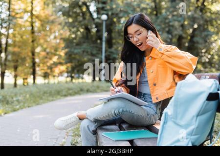 femme asiatique stupéfait d'écrire dans un ordinateur portable tout en parlant sur un smartphone sur un banc dans le parc Banque D'Images