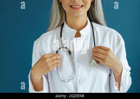 Sourire mature médecin en robe blanche avec stéthoscope se tient sur fond bleu Banque D'Images