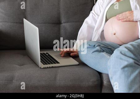 Jeune femme enceinte attrayante travaillant à distance en ligne depuis la maison. Gros plan femme mains sur ordinateur portable avec grand ventre avancé grossesse. Banque D'Images