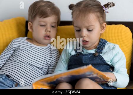 Deux enfants avant-garçonnets lisent un grand livre intéressant de contes de fées sur un lit jaune. Frères et sœurs petits jumeaux lecteur ont plaisir, heureux enfant sur Banque D'Images