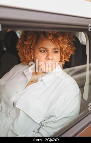 Joyeux jeune femme afro-américaine souriante rouge afro a aimé le conducteur assis dans une nouvelle voiture brune, souriant regardant la caméra appréciant le voyage. Cours de conduite et concept d'assurance vie. Banque D'Images