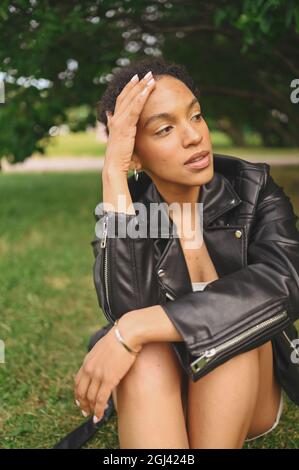 Portrait de mode de beau jeune sûr et attrayant naturel afro-américaine avec cheveux afro en cuir blouson noir posant dans le parc naturel en feuillage vert. Banque D'Images