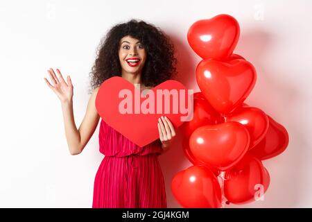 Concept Saint Valentin et amour. Jeune femme gaie dans une elegante robe rouge debout pres de ballons romantiques et tenant de gros coeur rouge decoupe agitant Photo Stock Alamy