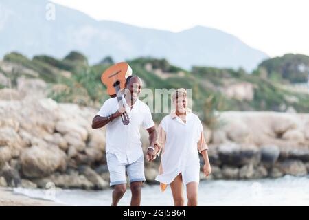 Bonne course mixte, couple d'âge moyen embrassant tout en marchant sur la plage tenant une guitare. Homme tenant guitare avec femme. Banque D'Images