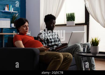 Couple interracial s'attendant à l'enfant et utilisant la technologie à la maison. Homme afro-américain travaillant sur un ordinateur portable numérique alors qu'une femme blanche enceinte pose sur un canapé. La race mixte partenaires avec la grossesse Banque D'Images