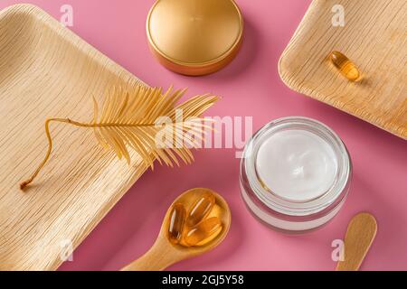 Crème blanche pour le soin de la peau dans un pot en verre ouvert, quelques capsules d'huile essentielle dans une cuillère en bois et feuille sèche jaune doré sur fond rose pastel. Banque D'Images