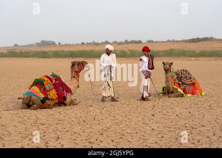 Inde, Gujarat, Bhuj, Grand Rann de Kutch, Tribe. Deux hommes se tiennent avec leurs chameaux dans le désert tandis que l'un fume un tuyau. (Usage éditorial uniquement) Banque D'Images