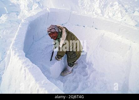 Baker Lake, Nunavut, Canada. Homme aîné inuit, habillé de vêtements modernes d'hiver, coupant des blocs de glace avec un couteau et les tailler tout en construisant le commerce Banque D'Images