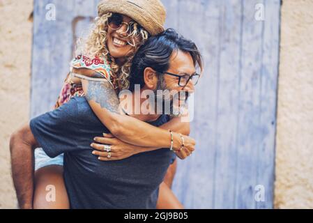 Couple insouciant passant du temps libre ensemble à l'extérieur, homme donnant pigegyback tour à la femme joyeuse tatouée dans des lunettes de soleil et chapeau de paille. Femme aime Banque D'Images