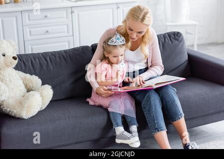 femme pointant vers le livre tout en lisant le conte de fées à la fille dans la couronne de jouet Banque D'Images