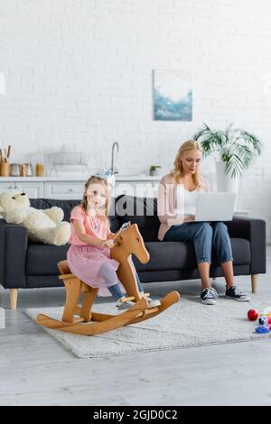 enfant dans la couronne jouet cheval à bascule près de maman floue travaillant sur ordinateur portable Banque D'Images