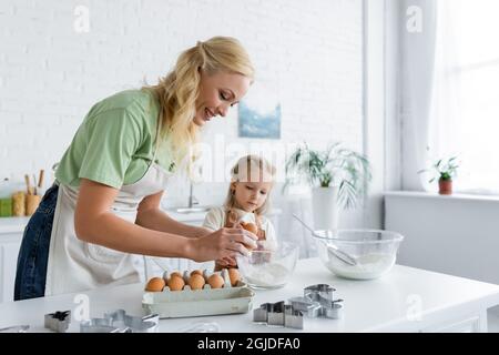 femme cassant l'œuf dans le bol tout en cuisinant avec la fille dans la cuisine Banque D'Images