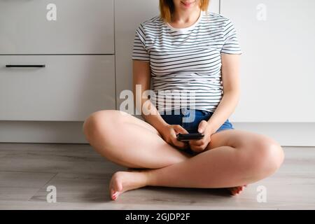 La jeune femme utilise un smartphone assis sur le sol dans la cuisine moderne. Femme envoyant un message texte avec son petit ami. Indépendant travaillant à domicile Banque D'Images