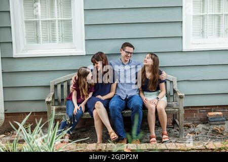 Une famille de quatre personnes avec une mère et un père et deux filles assises sur un banc à l'extérieur d'une maison de campagne bleue Banque D'Images