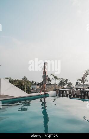 Un jeune homme de race blanche marche le long du bord de la piscine sur le toit de l'hôtel, donnant sur les palmiers. Eau cristalline Banque D'Images