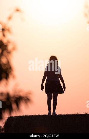 silhouette d'une femme dans le champ sur une balle de paille au coucher du soleil avec le soleil derrière elle et branches d'arbre sur les côtés. Image chaude. Banque D'Images