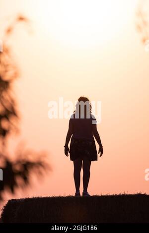 silhouette d'une femme dans le champ sur une balle de paille au coucher du soleil avec le soleil derrière elle et branches d'arbre sur les côtés. Image chaude. Banque D'Images