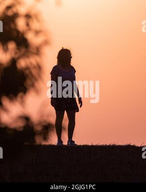 silhouette d'une femme dans le champ sur une balle de paille au coucher du soleil avec le soleil derrière elle et branches d'arbre sur les côtés. Image chaude. Banque D'Images