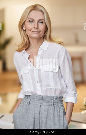 Confiant et réussi. Belle femme mûre avec des cheveux blonds regardant la caméra avec le sourire en posant à l'intérieur Banque D'Images