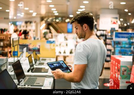 Vue latérale d'un jeune homme utilisant une tablette tout en recherchant un ordinateur portable numérique dans un magasin moderne avec divers gadgets Banque D'Images