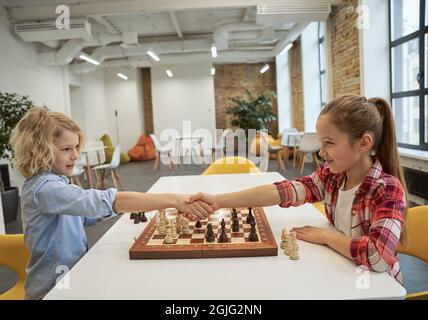 Merci. Un garçon et une fille caucasiens se regardant les uns les autres et se serrant la main après avoir joué aux échecs, assis ensemble à la table à l'école Banque D'Images