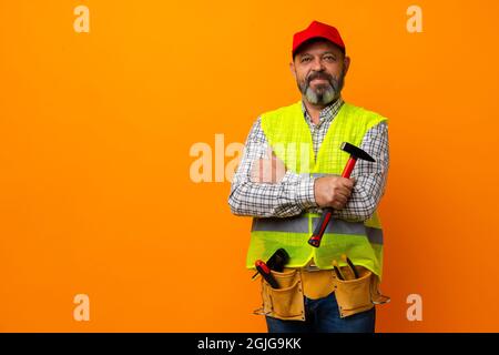 Homme barbu d'âge moyen constructeur en uniforme et casque avec outils sur fond orange Banque D'Images