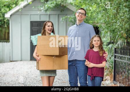 Un père et ses deux filles tenant des boîtes mobiles à l'extérieur sur une allée de gravier près d'un bâtiment d'entreposage Banque D'Images