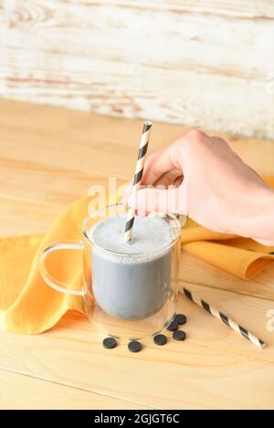 Femme mettant de la paille dans une tasse de verre de délicieux Latte au charbon de bois sur fond de bois Banque D'Images